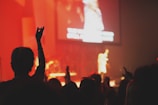 A concert scene with a silhouette of a crowd in the foreground and a performer on stage under bright lights. The atmosphere is lively with raised hands, indicating engagement and enthusiasm. The backdrop glows with warm, red tones.