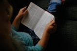 A child wearing a blue sweater and jeans is reading a book with closely spaced text while sitting on a dark patterned fabric sofa. The focus is on the open book held in the child's hands.