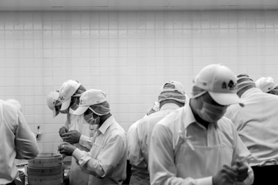 A group of people wearing white uniforms, caps, and masks are focused on a task in what appears to be a kitchen or food preparation area. The background features a tiled wall and there are stacked bamboo steamers on a counter.
