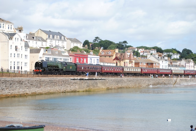 A steam locomotive with vintage-style passenger carriages travels along a coastal railway beside a stone seawall. Behind the train, a row of colorful houses with red, blue, and white facades is situated on a slight hill, with greenery and trees in the background. The scene is set against a partly cloudy sky, with a calm water body in the foreground.