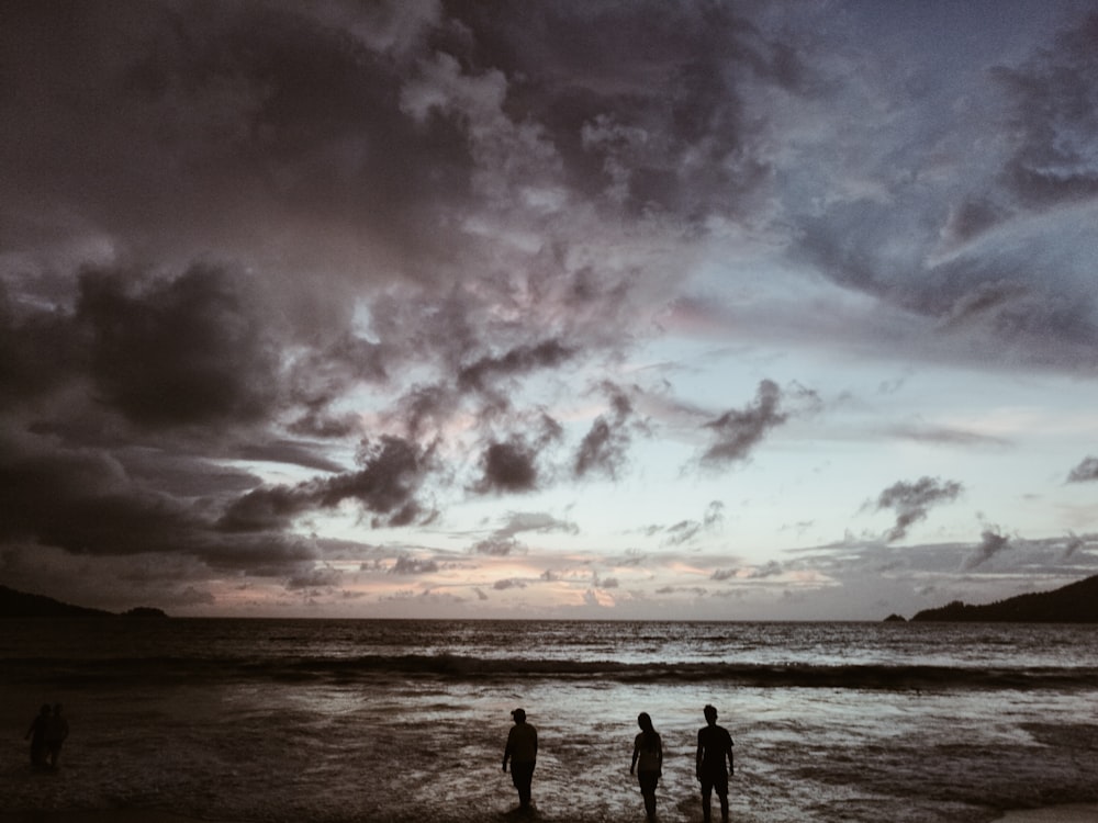 Silhouette von drei Menschen am Meeresufer unter grauem, bewölktem Himmel