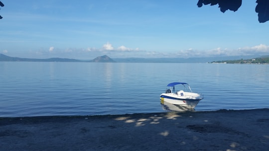 A small motorboat with a blue canopy floats on a calm lake with a distant volcano and hills in the background. The shoreline has sandy terrain with shadows of overhanging leaves.