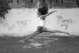 A person is captured mid-air in a graceful, dynamic leap against a graffiti-covered wall. The scene is outdoors, featuring a patch of grass and a background of concrete. The person's hair and clothing suggest movement and freedom, creating a striking visual against the urban backdrop.