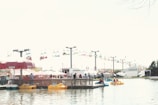 A busy amusement park scene with colorful chairlifts in the background. People enjoy paddle boating on a lake under clear skies. Bright vendor tents and a large restroom trailer are visible near the water’s edge.