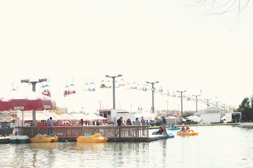 A busy amusement park scene with colorful chairlifts in the background. People enjoy paddle boating on a lake under clear skies. Bright vendor tents and a large restroom trailer are visible near the water’s edge.