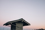 A wooden shelter with a green roof is depicted against a pastel-colored sky. The structure features exposed beams and support braces, standing solitary in an open area. The sky transitions in color from soft pink to blue, evoking a peaceful dusk or dawn setting. In the background, there is a hint of industrial structures and trees.