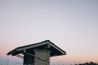 A wooden shelter with a green roof is depicted against a pastel-colored sky. The structure features exposed beams and support braces, standing solitary in an open area. The sky transitions in color from soft pink to blue, evoking a peaceful dusk or dawn setting. In the background, there is a hint of industrial structures and trees.