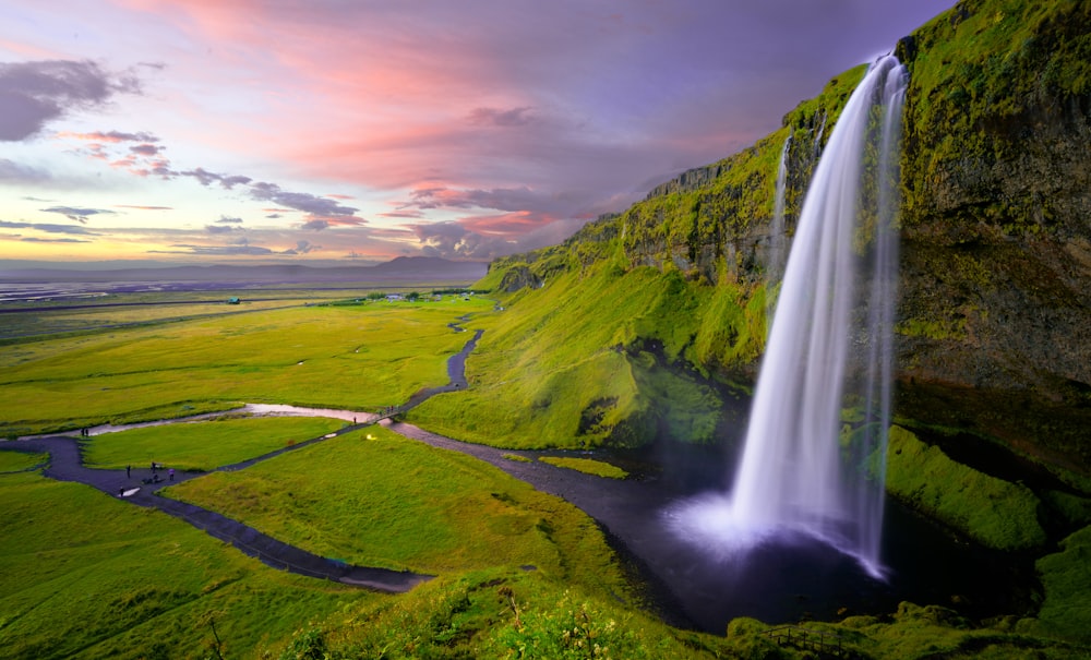 A beautiful landscape with green plains and a tall waterfall pouring from a moss-covered rock face