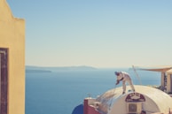 A person is painting the rooftop of a building with a stunning view of the ocean in the background. The building has a dome-shaped roof with a sign that says 'Maria Baba Vida'. The sea's blue waters meet the clear sky on the horizon, and there are hints of distant landforms.
