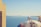 A person is painting the rooftop of a building with a stunning view of the ocean in the background. The building has a dome-shaped roof with a sign that says 'Maria Baba Vida'. The sea's blue waters meet the clear sky on the horizon, and there are hints of distant landforms.