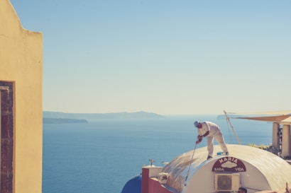 A person is painting the rooftop of a building with a stunning view of the ocean in the background. The building has a dome-shaped roof with a sign that says 'Maria Baba Vida'. The sea's blue waters meet the clear sky on the horizon, and there are hints of distant landforms.