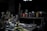 A dimly lit artist's studio workspace filled with various painting supplies. Brushes of different sizes are clustered in containers, surrounded by jars of paint in multiple colors. A table is covered with a textured cloth, and a selection of palette knives and paint tubes are scattered on it. Shelves hold additional paint jars and art tools, creating an atmosphere of creativity and artistic exploration.