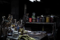 A dimly lit artist's studio workspace filled with various painting supplies. Brushes of different sizes are clustered in containers, surrounded by jars of paint in multiple colors. A table is covered with a textured cloth, and a selection of palette knives and paint tubes are scattered on it. Shelves hold additional paint jars and art tools, creating an atmosphere of creativity and artistic exploration.