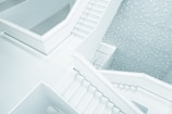 A modern architectural interior featuring multiple intersecting staircases with minimalist design. The surfaces are smooth and predominantly white, complemented by decorative patterns on the walls, creating a clean and contemporary aesthetic.