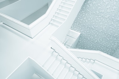 A modern architectural interior featuring multiple intersecting staircases with minimalist design. The surfaces are smooth and predominantly white, complemented by decorative patterns on the walls, creating a clean and contemporary aesthetic.
