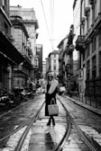 A woman wearing a long coat with a fur collar stands confidently on a wet cobblestone street, which has tram tracks running along its length. She is holding a stylish handbag and wearing sunglasses. The street is lined with classic European architecture, and several parked motorcycles are visible along the side.