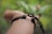 A close-up of a person's arm wearing a simple bracelet made of black and brown cords. The background is blurred, showing greenery, suggesting an outdoor setting.