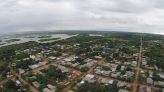 Aerial view of a rural town surrounded by lush greenery. The town has various buildings with tin roofs and dirt roads that intersect at right angles. In the background, expansive wetlands or lakes are visible, extending towards the horizon under a cloudy sky.