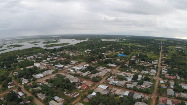 Aerial view of a rural town surrounded by lush greenery. The town has various buildings with tin roofs and dirt roads that intersect at right angles. In the background, expansive wetlands or lakes are visible, extending towards the horizon under a cloudy sky.