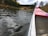 A scenic view of a calm lake surrounded by trees, with several people engaging in recreational water activities. Their reflections can be seen on the water's surface. In the foreground, the side of a canoe with red accents is visible, indicating a perspective from someone sitting inside.