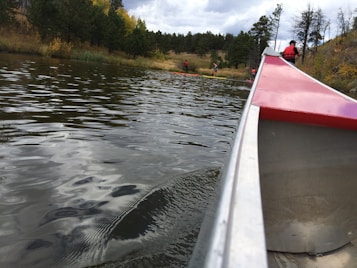 A scenic view of a calm lake surrounded by trees, with several people engaging in recreational water activities. Their reflections can be seen on the water's surface. In the foreground, the side of a canoe with red accents is visible, indicating a perspective from someone sitting inside.