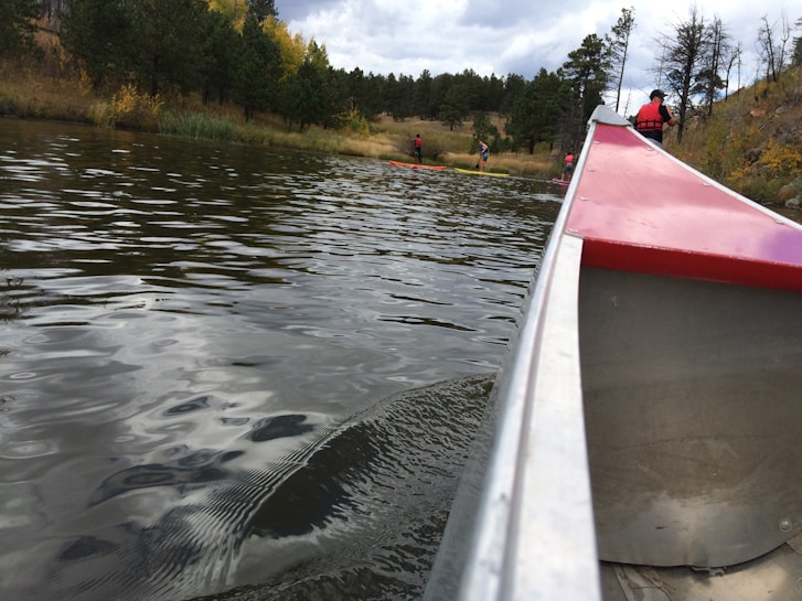 A scenic view of a calm lake surrounded by trees, with several people engaging in recreational water activities. Their reflections can be seen on the water's surface. In the foreground, the side of a canoe with red accents is visible, indicating a perspective from someone sitting inside.