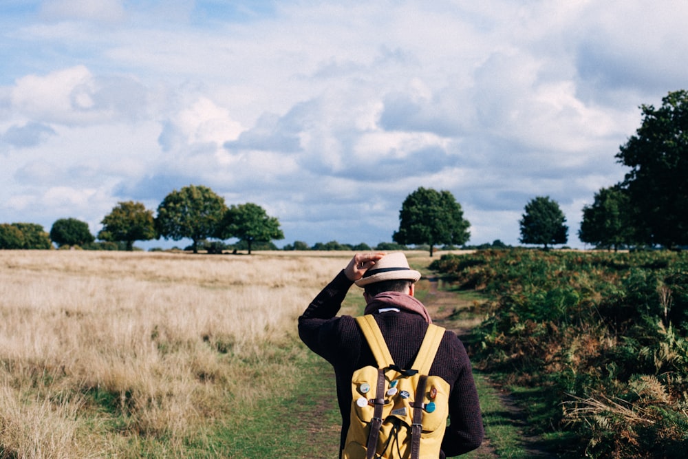 Hombre sosteniendo la gorra de fedora mientras camina sobre el campo de hierba verde y beige bajo nubes blancas