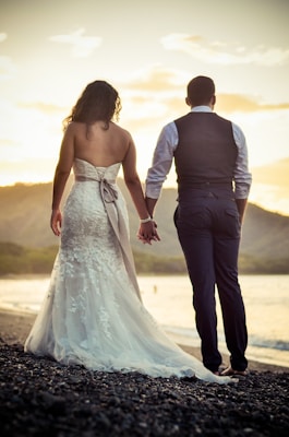 A couple stands hand in hand on a rocky beach, facing the ocean as the sun sets. The woman is wearing a white lace wedding dress with a long train, while the man is dressed in a dark suit with a vest and white shirt. The sky is filled with soft golden hues from the sunset, creating a warm atmosphere.