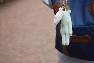 A plush toy with blue and white stripes and bear features hangs from a caramel-colored keychain attached to a blue backpack with brown leather accents. The backpack is labelled with the word 'OUTDOOR' and the setting is an outdoor area with a dirt path.