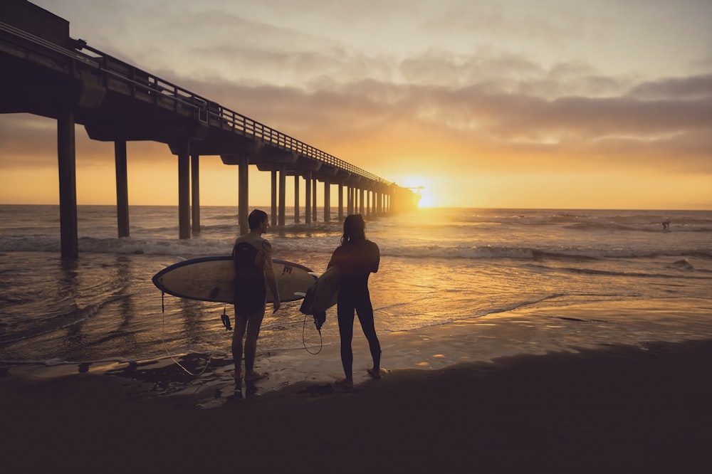 Frau trägt Surfbrett neben Person während des Sonnenuntergangs