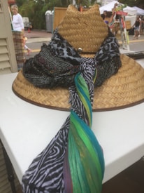 A woven straw hat is placed on a table, adorned with a variety of colorful and patterned scarves. The setting appears to be an outdoor market with people visible in the background. Display tents and a street can be seen, suggesting a lively and casual atmosphere.
