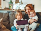 two babies and woman sitting on sofa while holding baby and watching on tablet