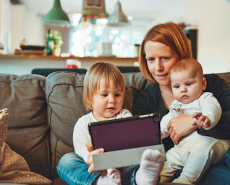 two babies and woman sitting on sofa while holding baby and watching on tablet