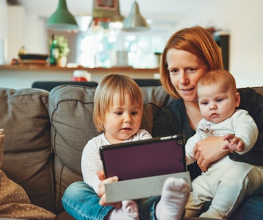 two babies and woman sitting on sofa while holding baby and watching on tablet