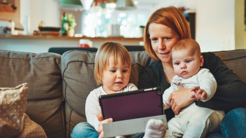 two babies and woman sitting on sofa while holding baby and watching on tablet