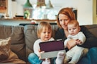 two babies and woman sitting on sofa while holding baby and watching on tablet