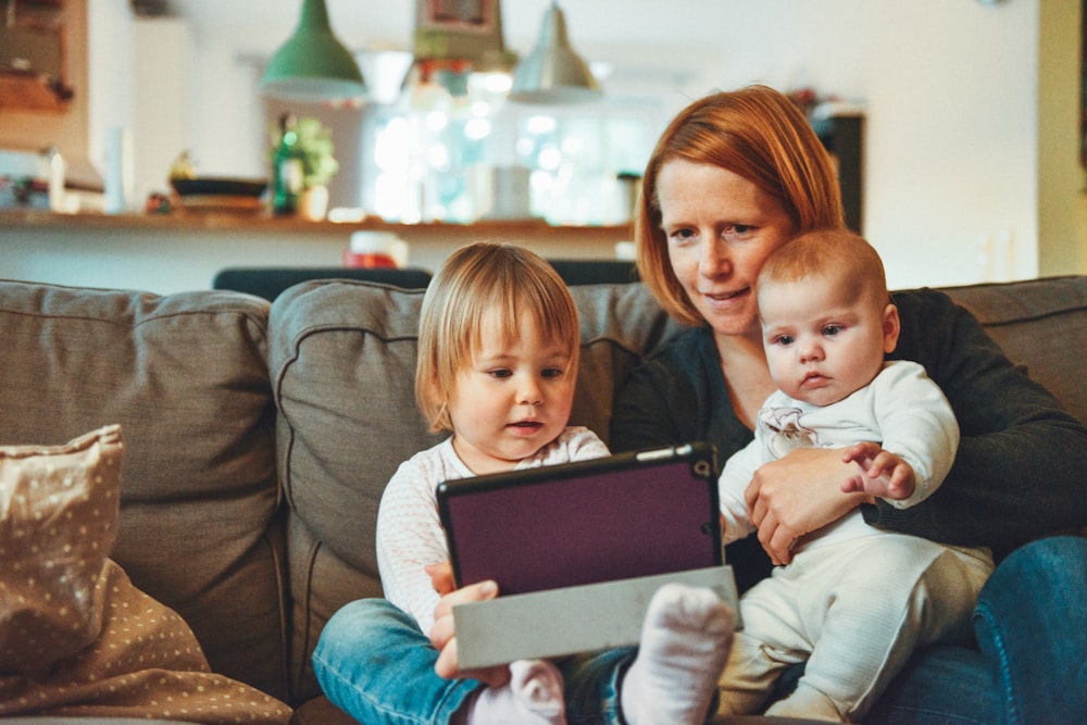 두 명의 아기와 여자가 아기를 안고 태블릿을 보면서 소파에 앉아 있습니다.