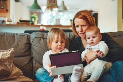 two babies and woman sitting on sofa while holding baby and watching on tablet parents google meet background