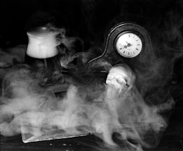 A dimly lit scene features a vintage clock with Roman numerals, a human skull, and a book partially obscured by swirling smoke. The objects give the setting an eerie and mysterious atmosphere.