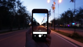 A hand holds a smartphone, capturing a long, empty road lined with trees under the evening sky. Surrounding streetlights are illuminated, casting a warm glow. The road stretches toward the horizon, creating a sense of depth.