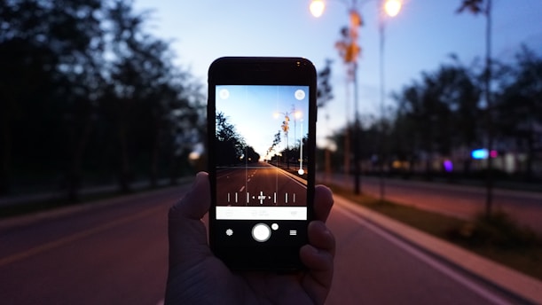 A hand holds a smartphone, capturing a long, empty road lined with trees under the evening sky. Surrounding streetlights are illuminated, casting a warm glow. The road stretches toward the horizon, creating a sense of depth.
