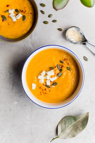 Bowls of orange soup topped with feta cheese and pumpkin seeds on a table