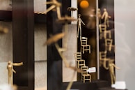 A display featuring artistic sculptures of stick figures made of metal. Some figures are climbing or balancing on small stacked chairs. One figure holds an umbrella, and there is a price tag visible.