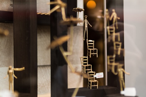 A display featuring artistic sculptures of stick figures made of metal. Some figures are climbing or balancing on small stacked chairs. One figure holds an umbrella, and there is a price tag visible.
