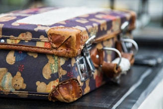 A vintage suitcase with worn leather corners and a playful teddy bear pattern on the fabric. The design showcases different colored bears against a dark background. The suitcase has metal latches and leather straps for closure.