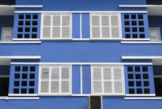 A vibrant blue building facade featuring symmetrical white shuttered windows. The architectural design includes a pattern of square openings surrounding the windows, creating a visually striking contrast between the blue background and the white elements.
