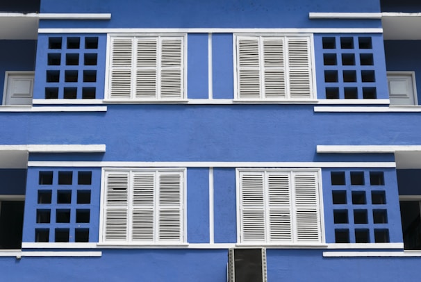 A vibrant blue building facade featuring symmetrical white shuttered windows. The architectural design includes a pattern of square openings surrounding the windows, creating a visually striking contrast between the blue background and the white elements.