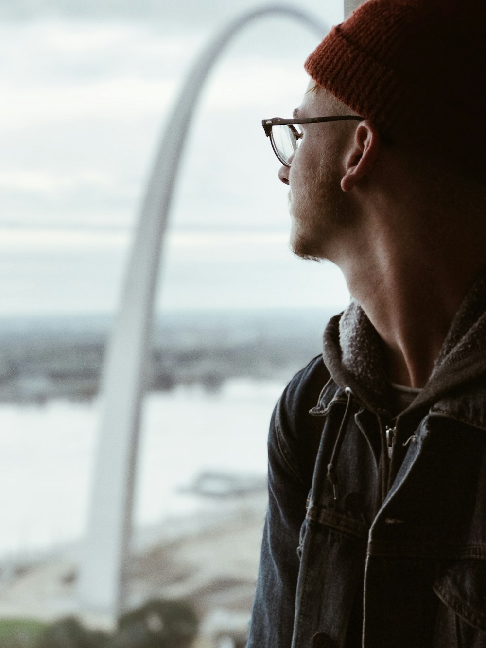 man in denim jacket standing beside window looking outside