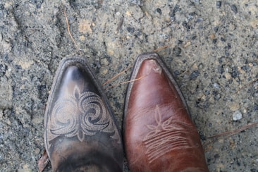 Two pointed-toe cowboy boots, one dark leather with intricate swirling patterns and the other brown with subtle embroidery, are shown on a rough, textured ground with small stones and patches of sand.