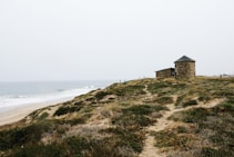 An expansive beach landscape with gentle waves approaching a sandy shore. A grassy dune with patches of green and brown vegetation leads to a small, cylindrical stone building with a conical roof situated on top of the hill. The sky is overcast, casting a muted light over the scene.